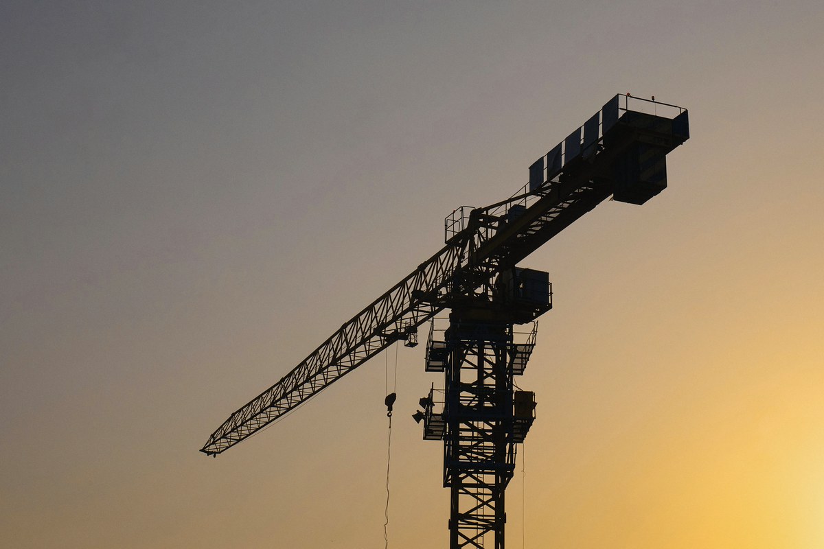 Silhouette of a tower crane, its jib angled down to the left, against a hazy dusk sky that fades from slate grey to warm gold