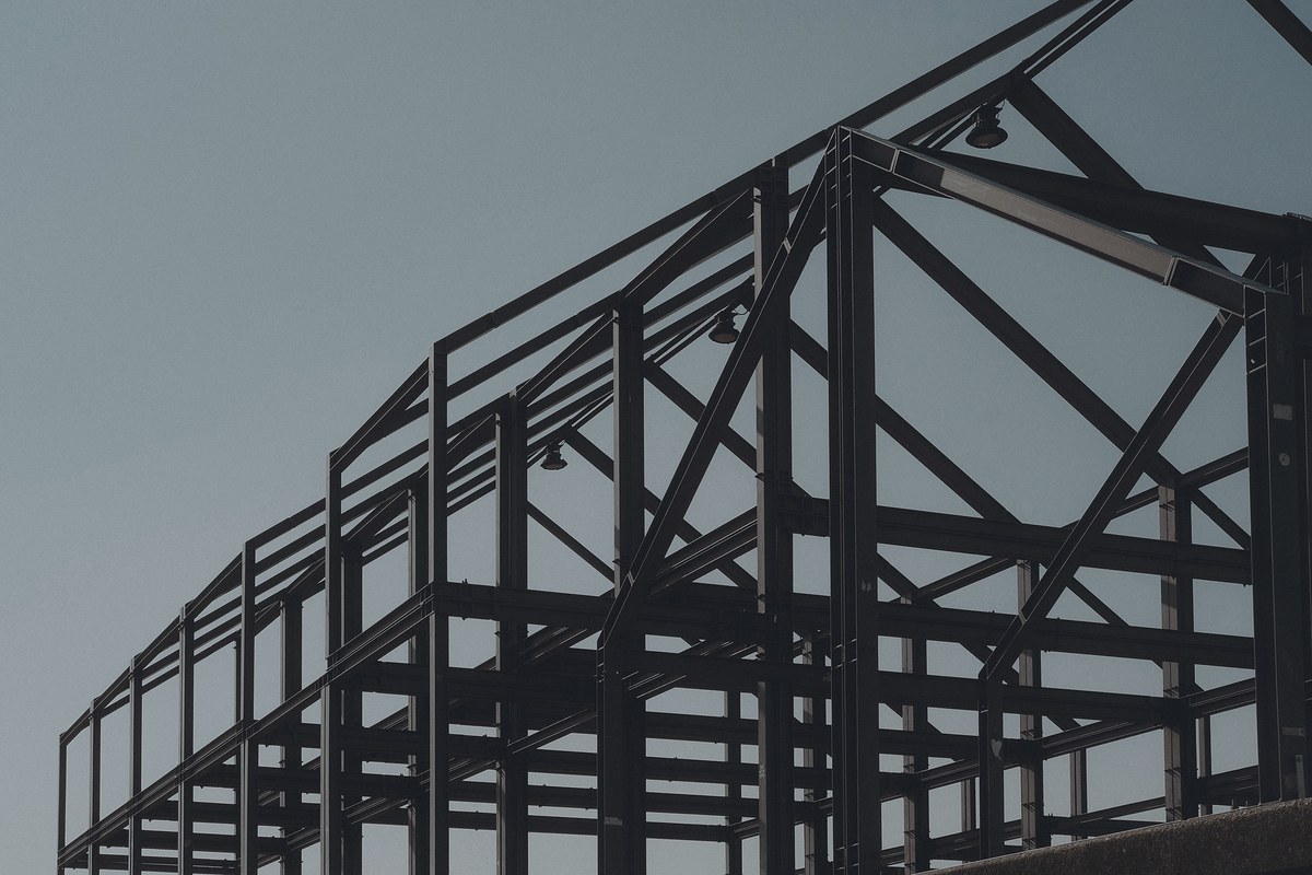 Dark steel frame of columns, beams and diagonal braces receding in rows against a flat pale blue-grey sky