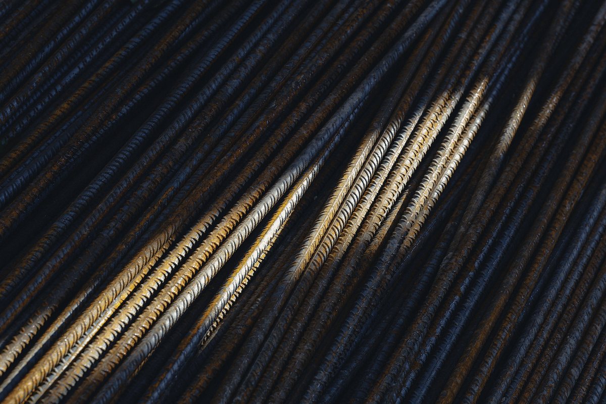 Close-up of ribbed steel reinforcing bars laid diagonally, mostly in deep shadow, with a band of warm sunlight picking out a few bars in gold