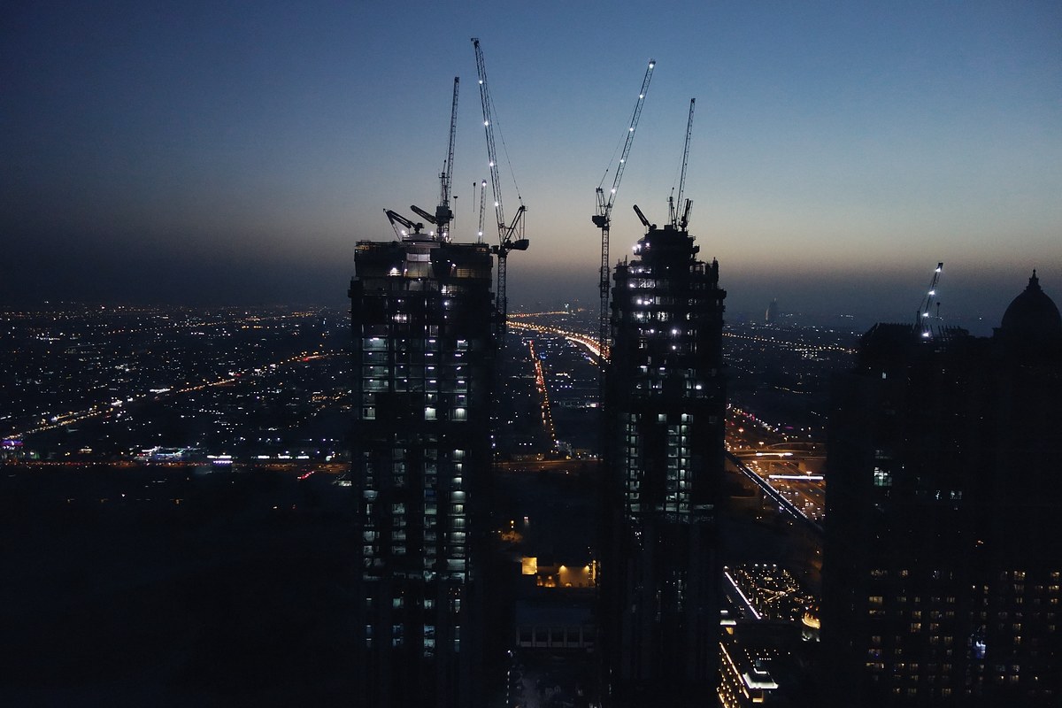 Two towers under construction at blue hour, topped by tower cranes strung with white work lights, above a dark city with lit roads stretching to the horizon