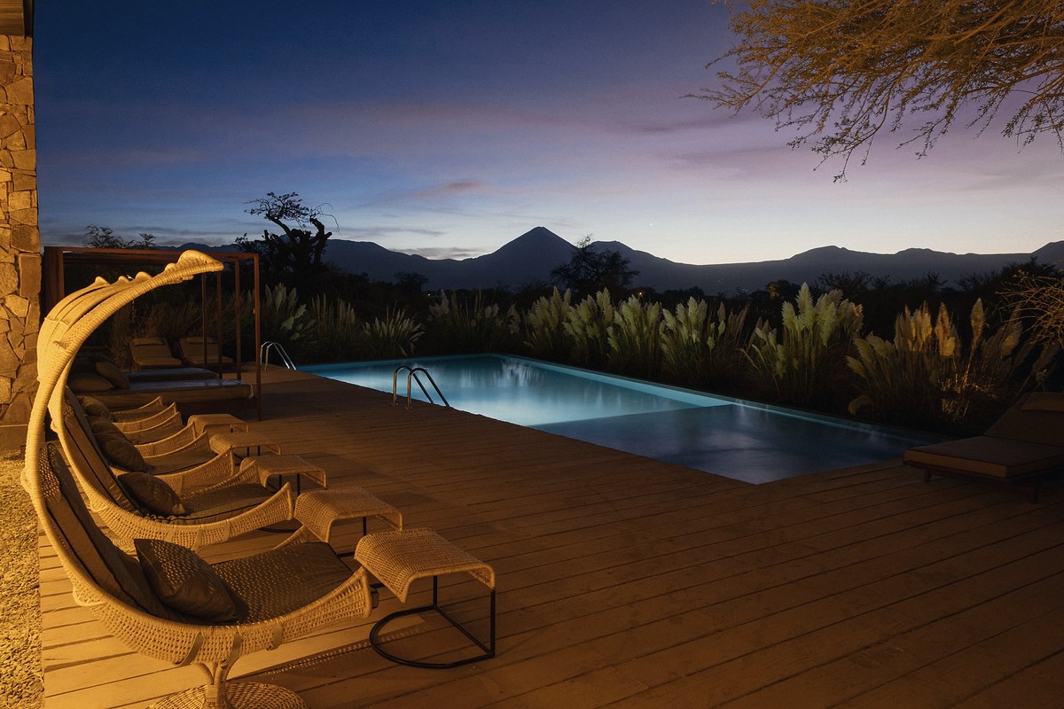 Lit swimming pool beside a timber deck at dusk, with woven loungers in warm lamplight on the left, pampas grass behind the water and a mountain ridge silhouetted under a deep blue and violet sky