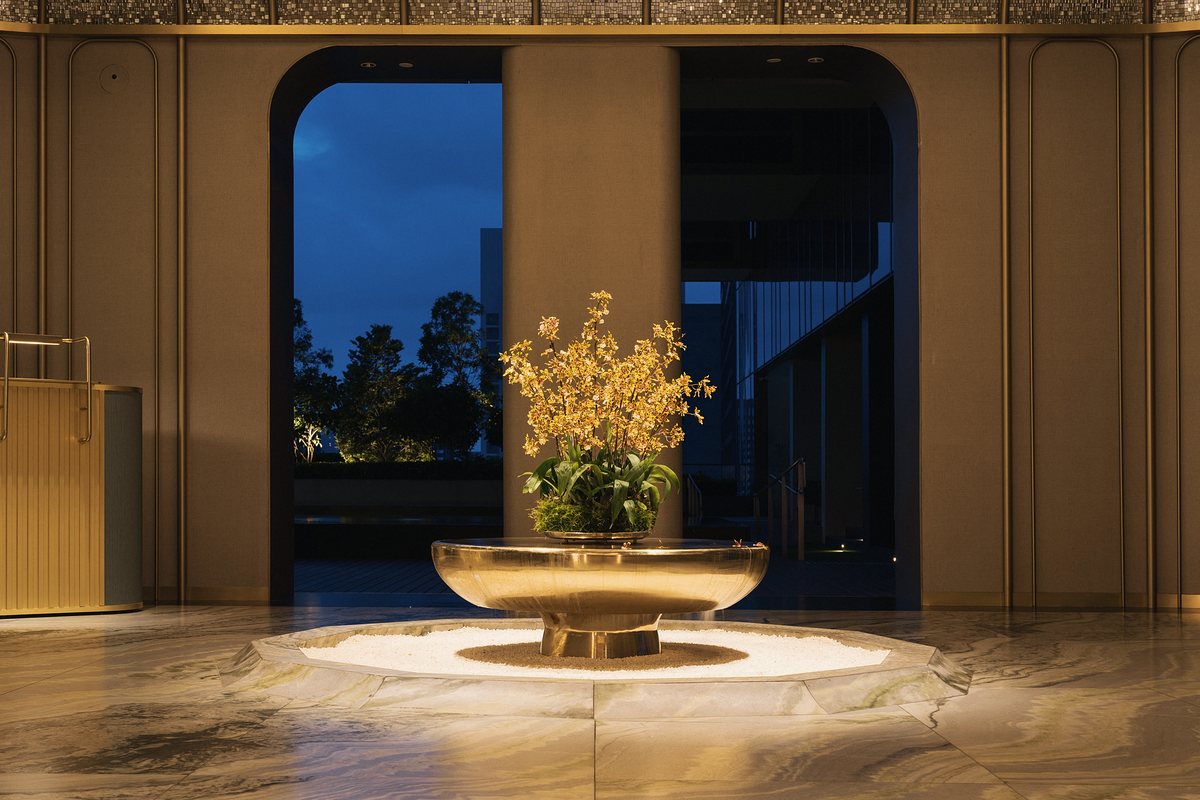 Polished brass bowl of yellow orchids on a lit ring of white pebbles set into a veined marble floor, in front of fabric wall panels with brass trim and arched openings onto a blue evening sky