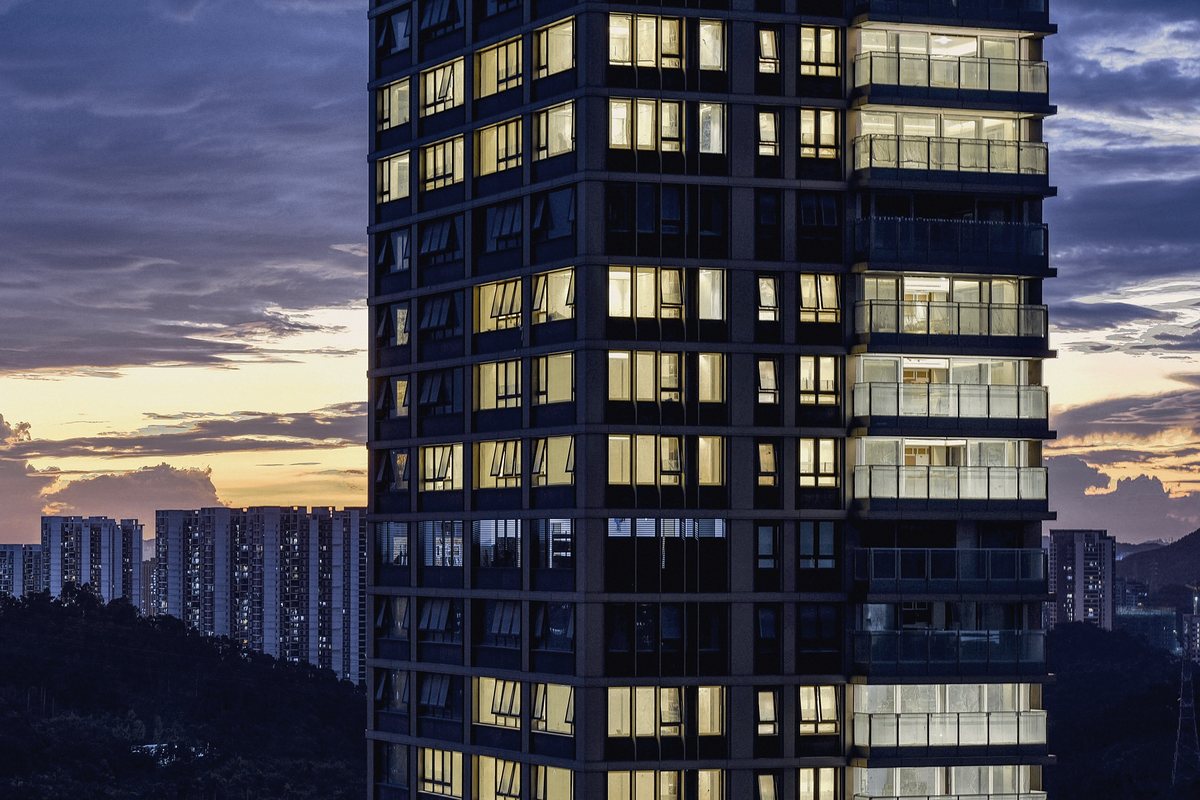 Dark residential tower with rows of warmly lit windows and glass balconies at dusk, distant apartment blocks and a wooded hill beneath a violet and amber sky