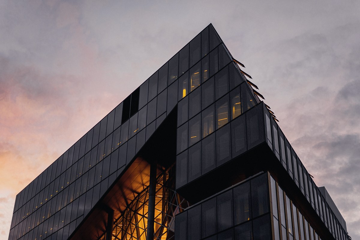 Low-angle view of an angular black glass building, its cut-away corner glowing amber from inside, against a soft pink and grey evening sky