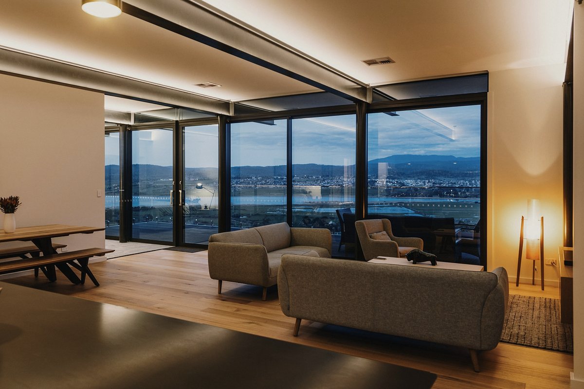 Open-plan living room at dusk with grey sofas, a timber dining table and floor-to-ceiling glass doors looking out over a bay and distant hills, lit by warm concealed ceiling lighting