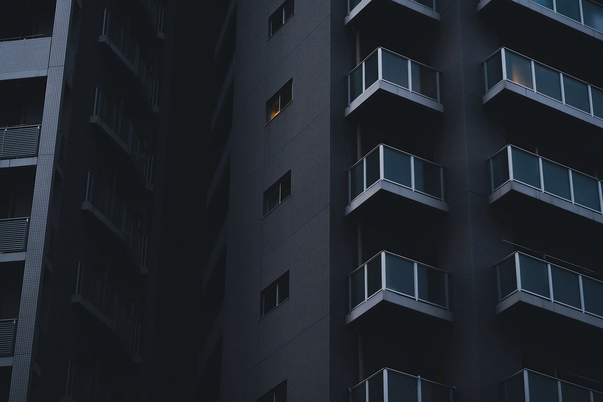 Close view of a dark apartment tower at dusk with stacked glass-fronted balconies on the right, a column of small windows with one lit amber, and railed balconies receding on the left